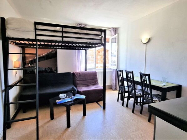 A compact room with a bunk bed, a sofa, a dining table with chairs, and a small table with a book.