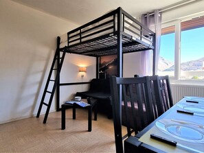 A bunk bed with a ladder, a table with cutlery, a chair, a small table with a bowl and a mug, and a wall-mounted lamp.