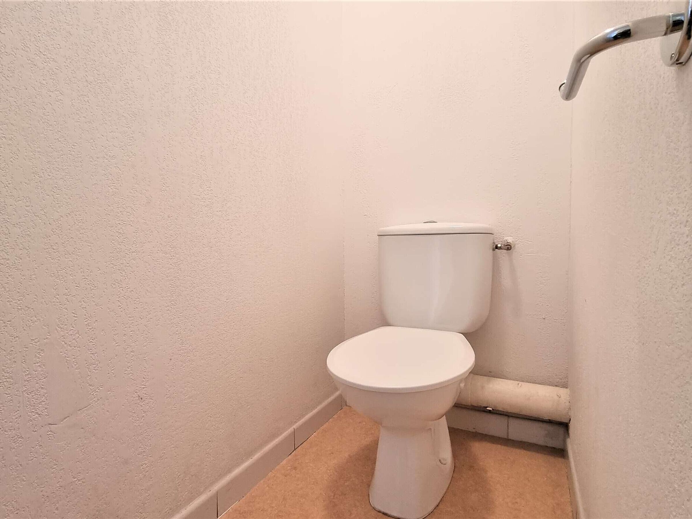 A small, white toilet in a corner of a room with plain walls and a beige floor.