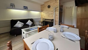 A dining area with a table set for four, a sofa with cushions, a wooden accent wall, and a television mounted on the wall. - Studio for 4 in SuperDévoluy with Wifi (Le Dévoluy)