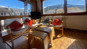 A sunlit room with a TV, two chairs with red cushions, a small table, and large windows offering a view of buildings and hills. - Family duplex in village center, pets allowed (Samoëns)