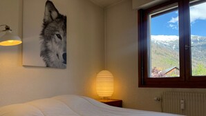 A bedroom with a bed, a wall-mounted lamp, a window with a view of mountains, and a wolf painting. - Family duplex in village center, pets allowed (Samoëns)