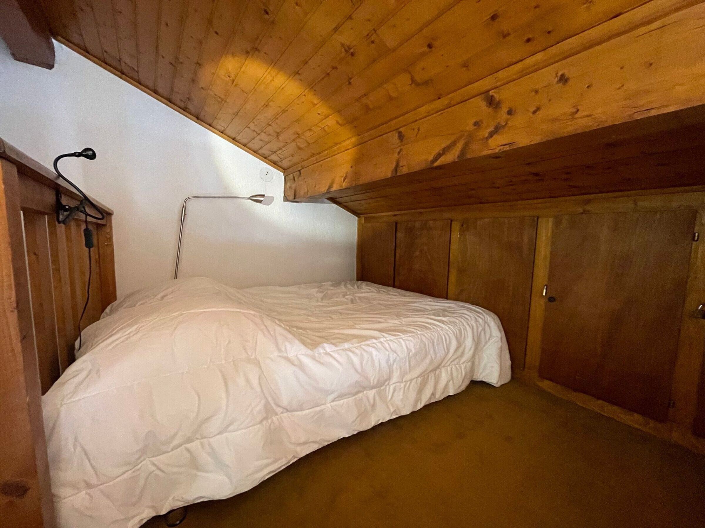 A single bed in a wooden room with a sloped ceiling, a wall-mounted light, and a door.