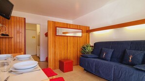 A room with a wooden wall, a blue sofa, a small red stool, a dining table set with plates and cutlery, and a television mounted on the wall.