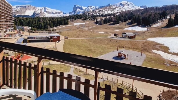 A mountain view from a balcony with a wooden railing, overlooking a ski resort area with buildings and snow-covered mountains.