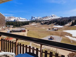 Une vue sur la montagne depuis un balcon doté d’une balustrade en bois, donnant sur une station de ski avec ses bâtiments et ses montagnes enneigées.