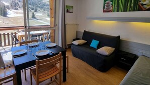 Un cuarto acogedor con una mesa de comedor dispuesta para dos personas, un sofá negro con cojines, un ventanal con vistas a un paisaje nevado y un estante de pared con objetos decorativos.
