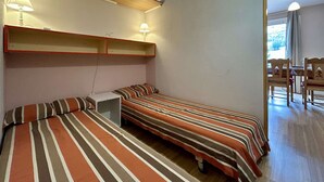 A dormitory room with two beds, a desk, and a chair. There are two wall-mounted lamps and a window providing natural light.