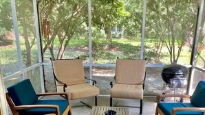 Une terrasse couverte, dotée de quatre chaises en bois et d’une table, et entourée de murs en verre offrant une vue sur un jardin.