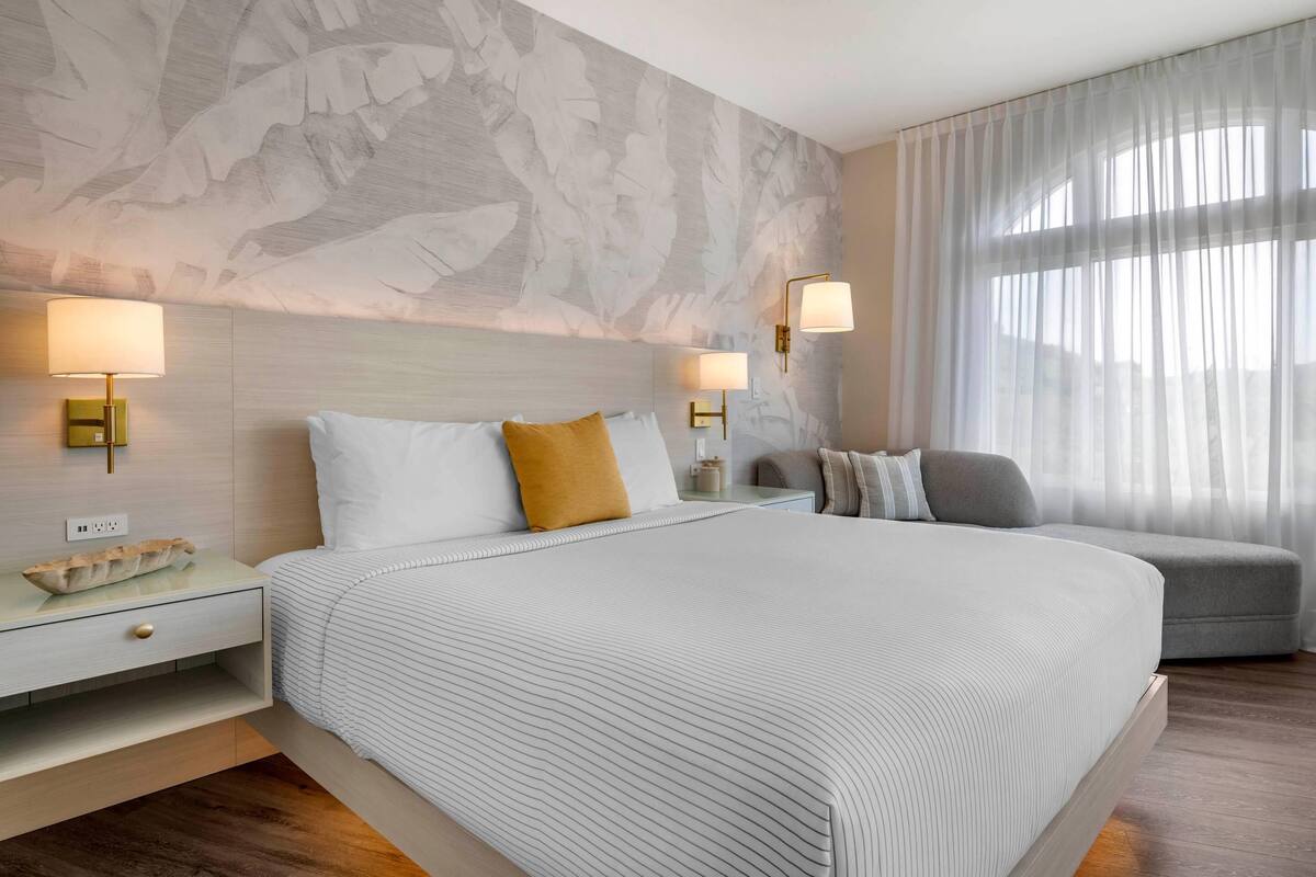 Habitación de hotel con una cama grande, dos mesitas de noche, una silla y una ventana con cortinas de gasa.