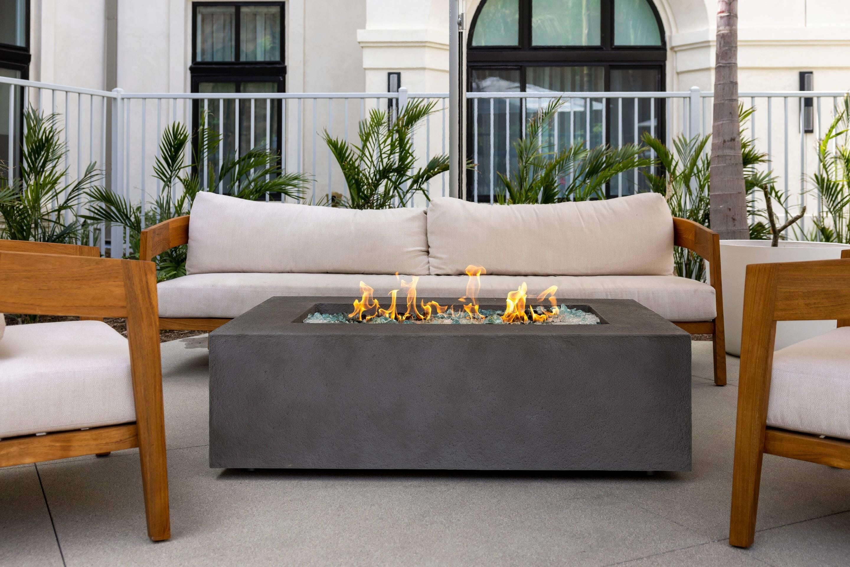 A modern outdoor seating area with a sofa and two wooden chairs, a square fire pit with flames, and a balcony with plants and a building in the background.