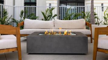 A modern outdoor seating area with a sofa and two wooden chairs, a square fire pit with flames, and a balcony with plants and a building in the background.
