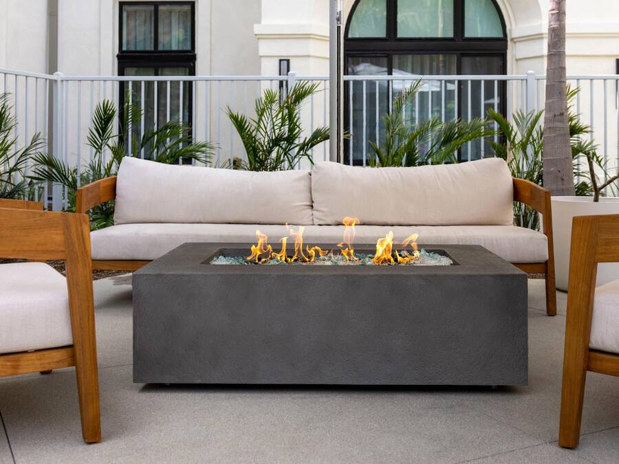 A modern outdoor seating area with a sofa and two wooden chairs, a square fire pit with flames, and a balcony with plants and a building in the background.