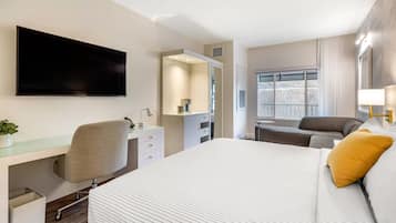 Quarto de hotel com uma cama grande, uma escrivaninha com cadeira, televisão na parede, um sofá e uma janela com cortinas.