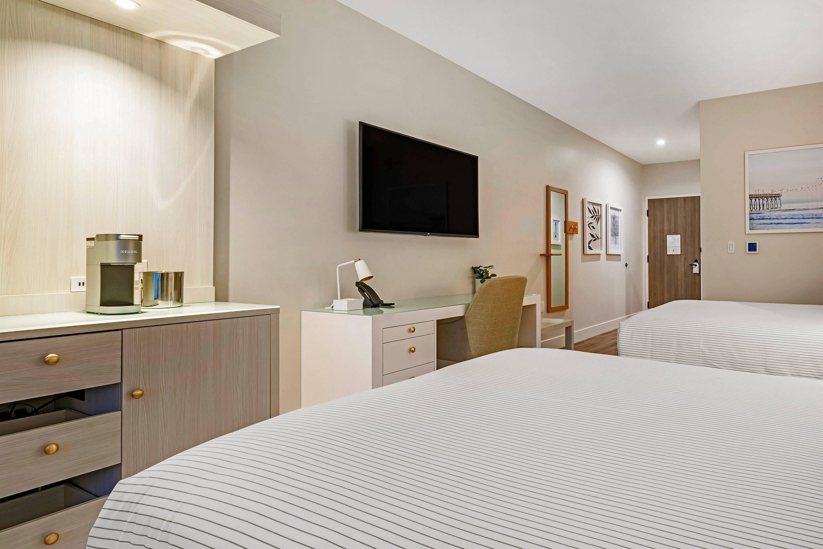 Habitación de hotel con dos camas, un escritorio con silla, un televisor de pantalla plana, cafetera y un cuadro en la pared.