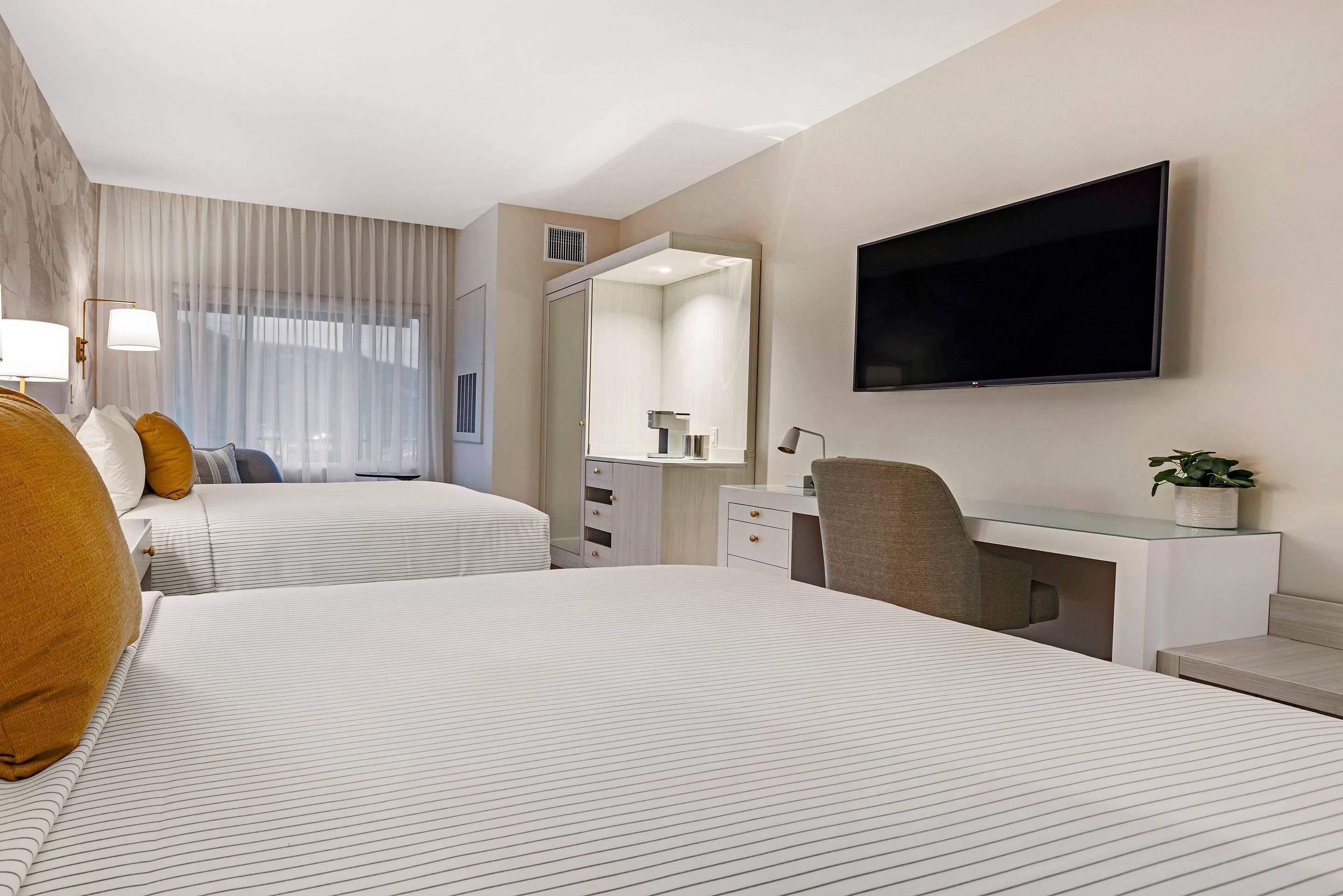 Ett hotellrum med två sängar, ett skrivbord, en stol, en TV och ett stort fönster med gardiner.