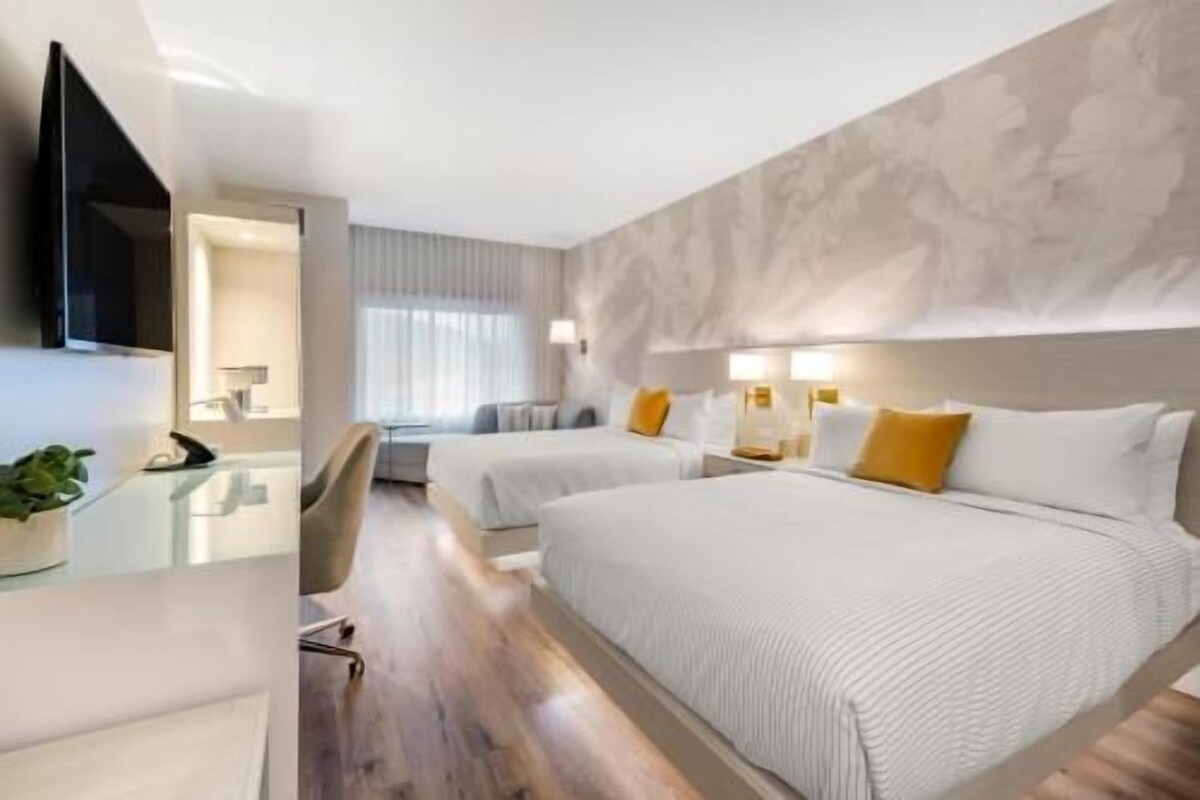 A hotel room with two beds, a flat-screen TV, a desk with a chair, and a large window with curtains.
