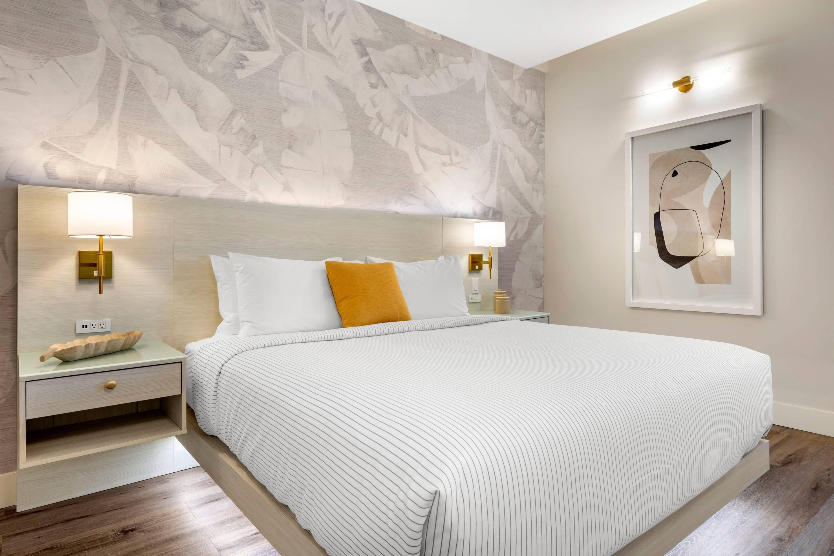 A modern hotel room with a large bed, two bedside tables with lamps, a wall mural, and a framed abstract artwork.