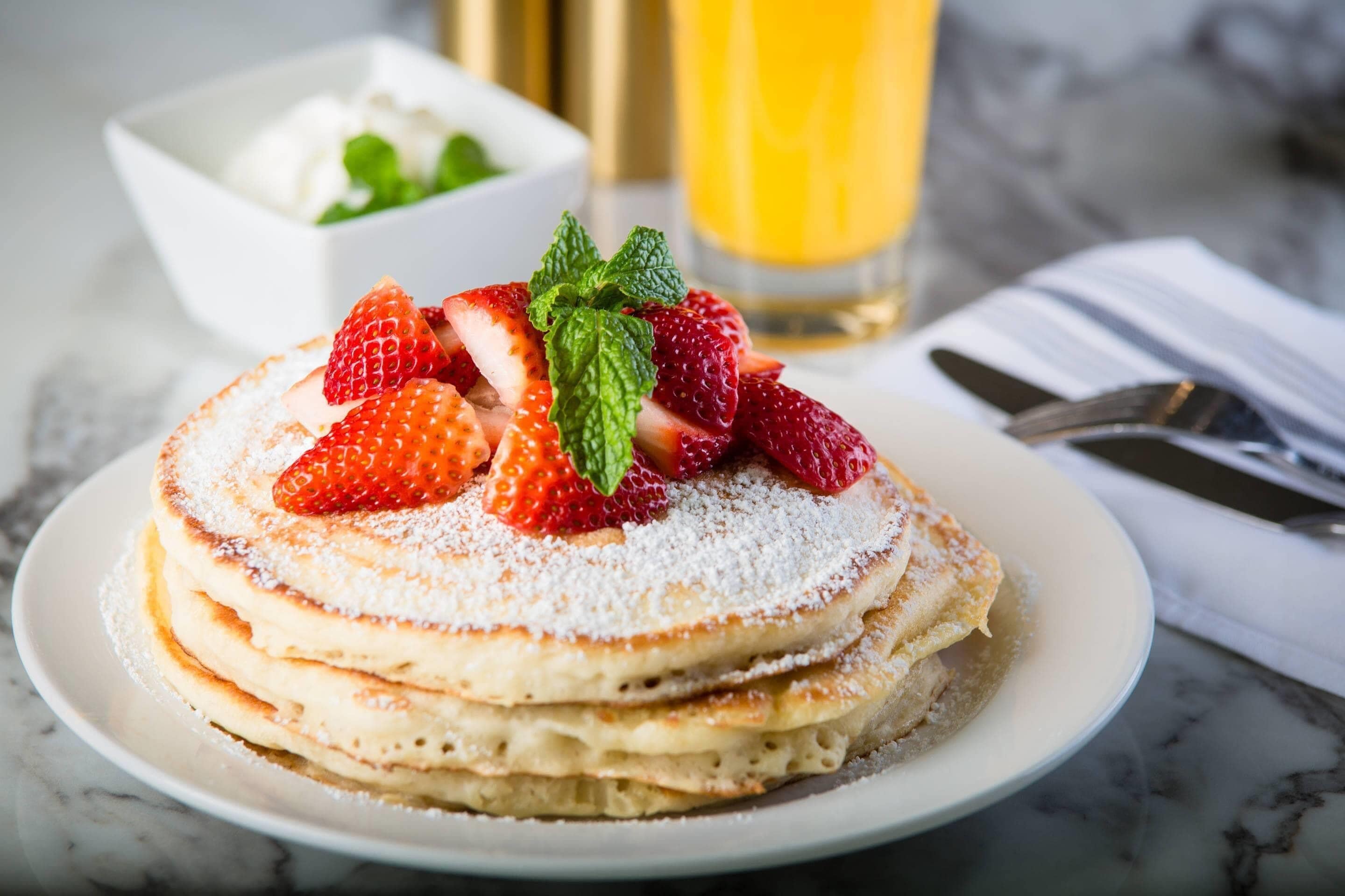 A stack of pancakes with strawberries and mint on a plate, with a glass of orange juice and a bowl of whipped cream in the background.