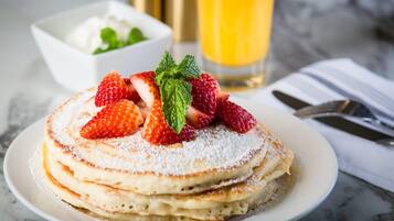 A stack of pancakes with strawberries and mint on a plate, with a glass of orange juice and a bowl of whipped cream in the background.