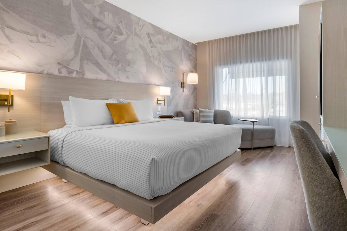 Una habitación de hotel moderna con una cama grande, mesitas de noche, una pequeña mesa, una silla y un ventanal con cortinas.