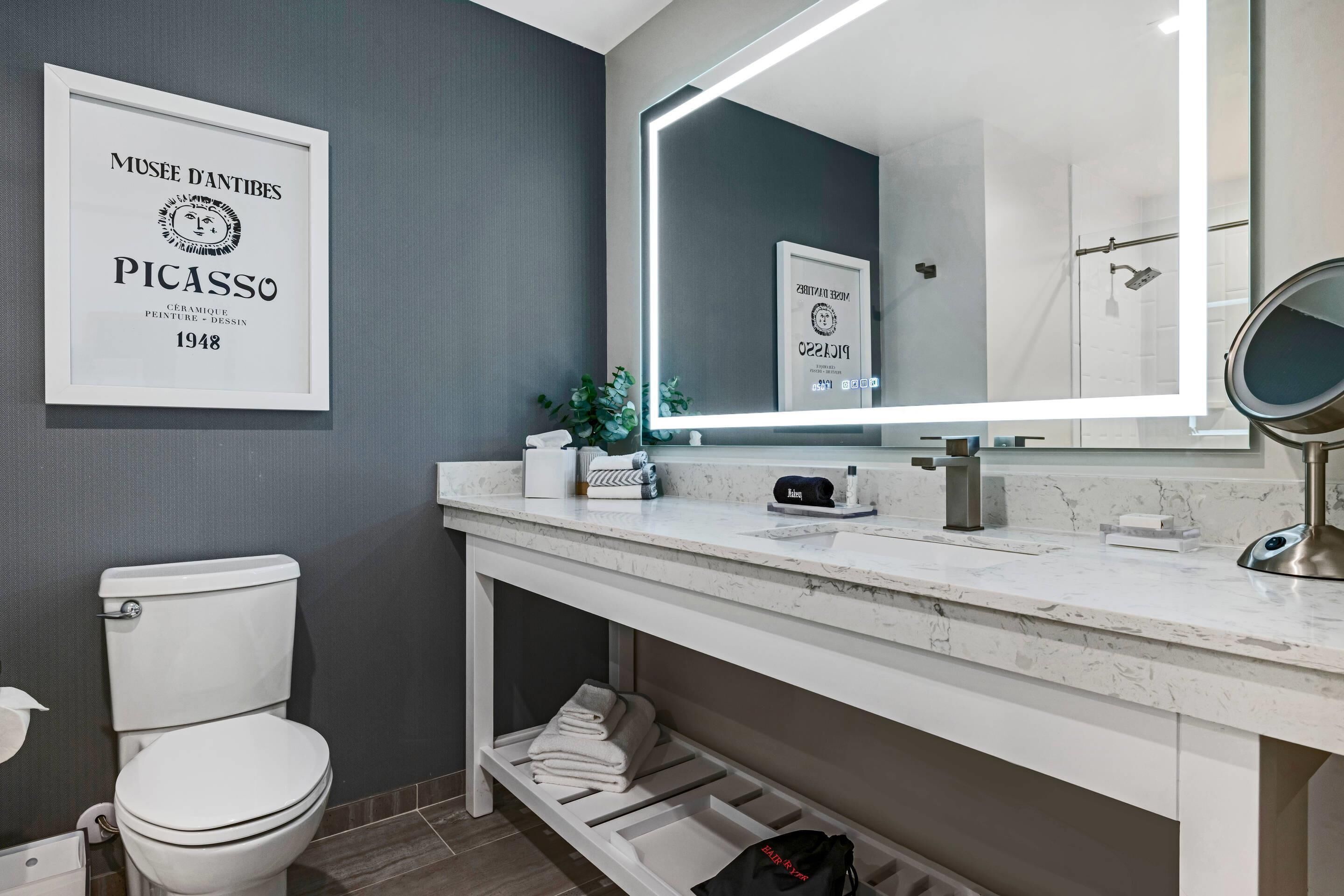 A modern bathroom with a toilet, sink, and mirror. There is a framed artwork on the wall and a shelf under the sink.