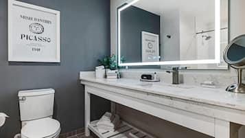 A modern bathroom with a toilet, sink, and mirror. There is a framed artwork on the wall and a shelf under the sink.