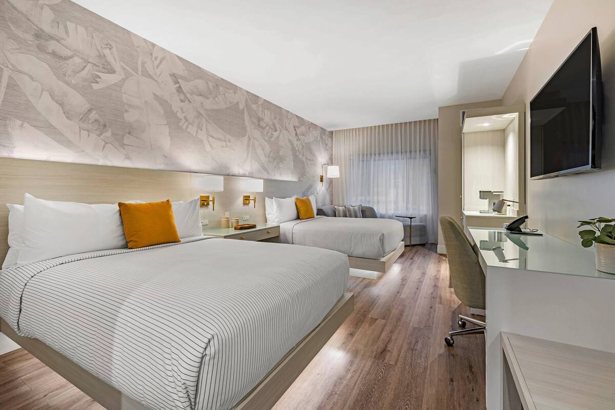 Habitación de hotel con dos camas, un escritorio, una silla, un televisor y un ventanal con cortinas.