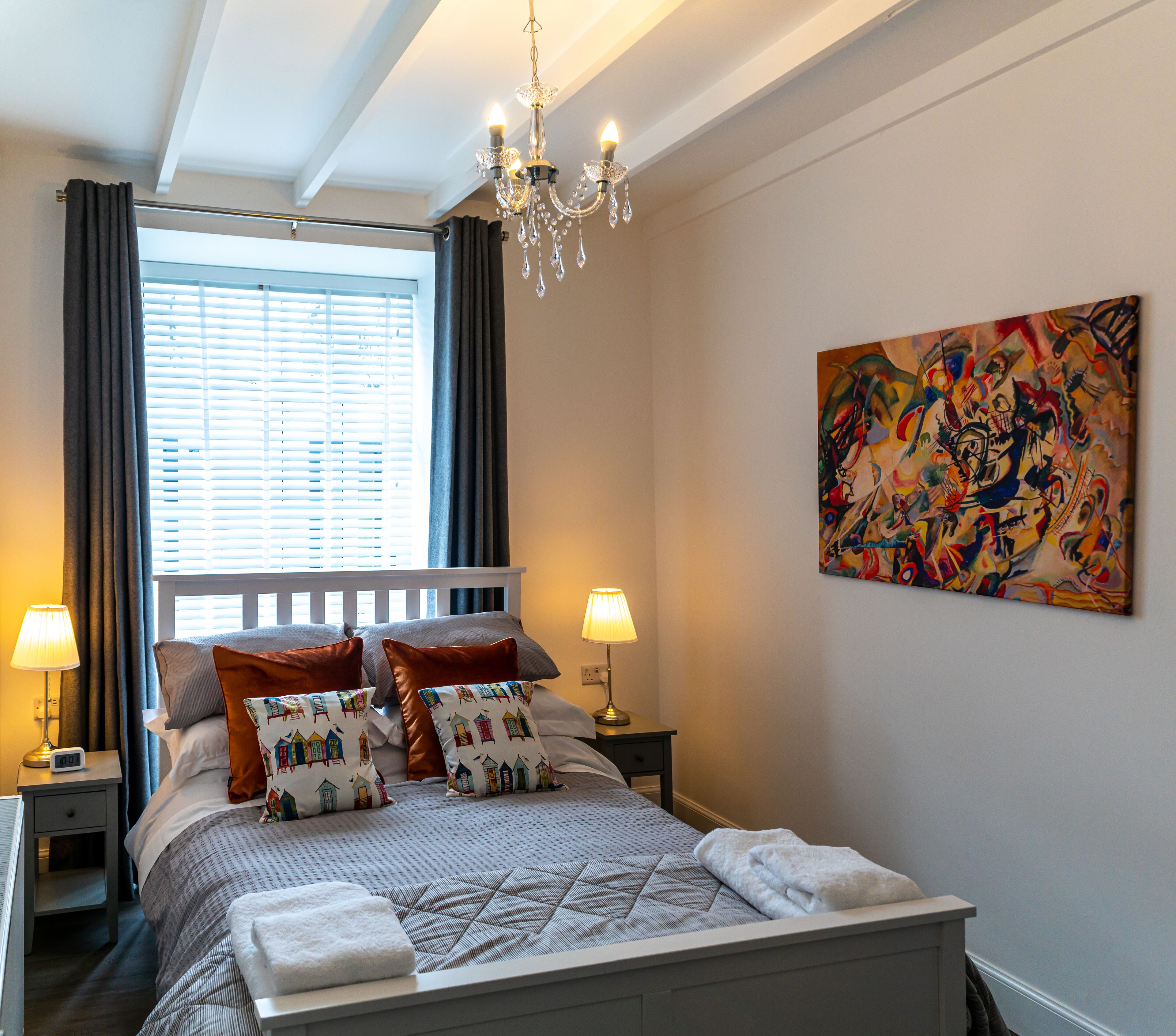 A neatly made bed with patterned pillows, a chandelier, a painting on the wall, and a window with curtains.