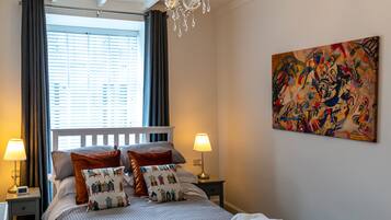 A neatly made bed with patterned pillows, a chandelier, a painting on the wall, and a window with curtains.