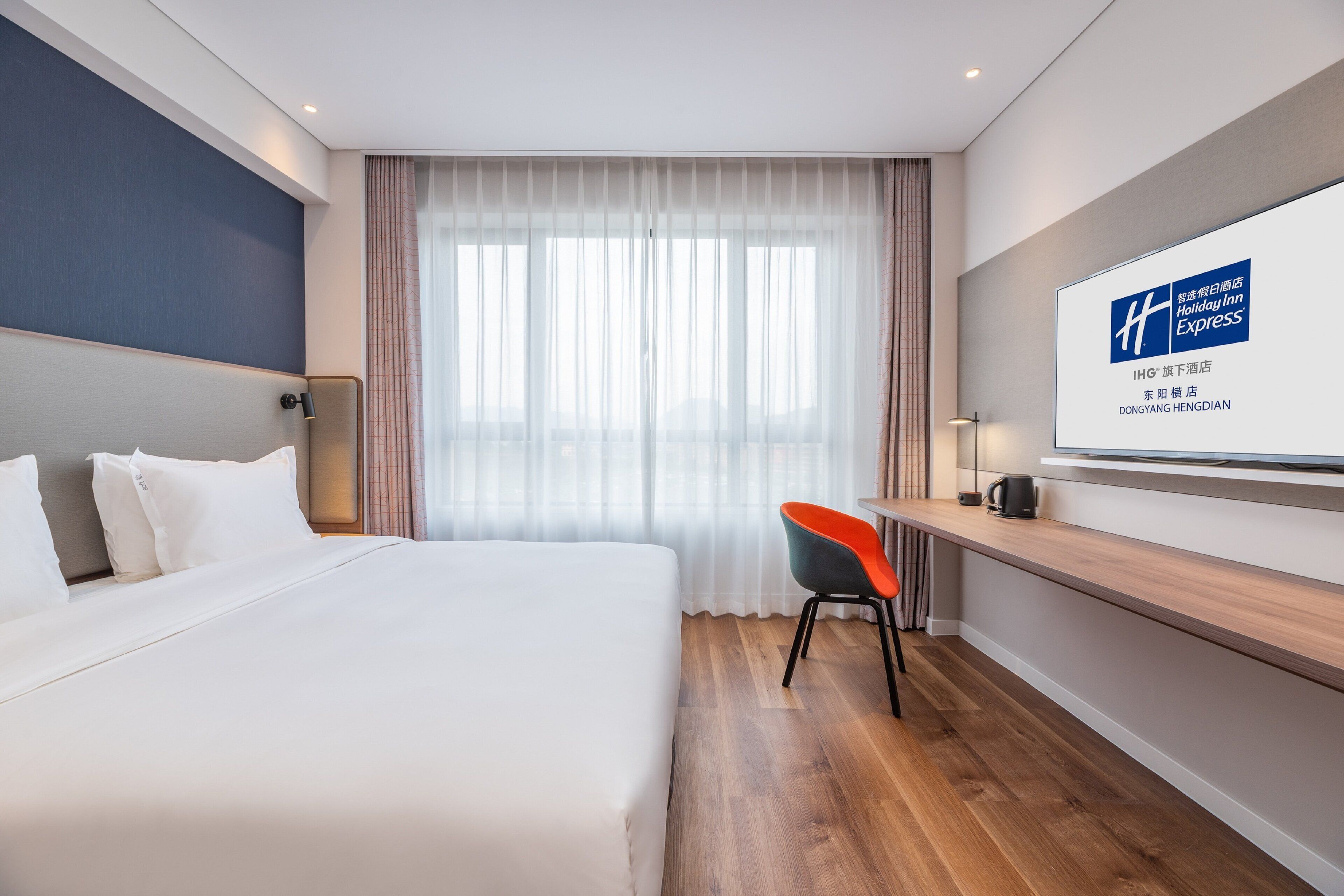 Hotellihuone, jossa on suuri sänky, työpöytä tuolilla sekä televisio, jossa näkyy hotellin nimi.