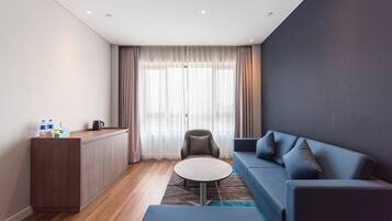 Una habitación de hotel moderna con piso de madera, un sofá, una mesa redonda, una silla, una pequeña área de cocina y un amplio ventanal con cortinas.