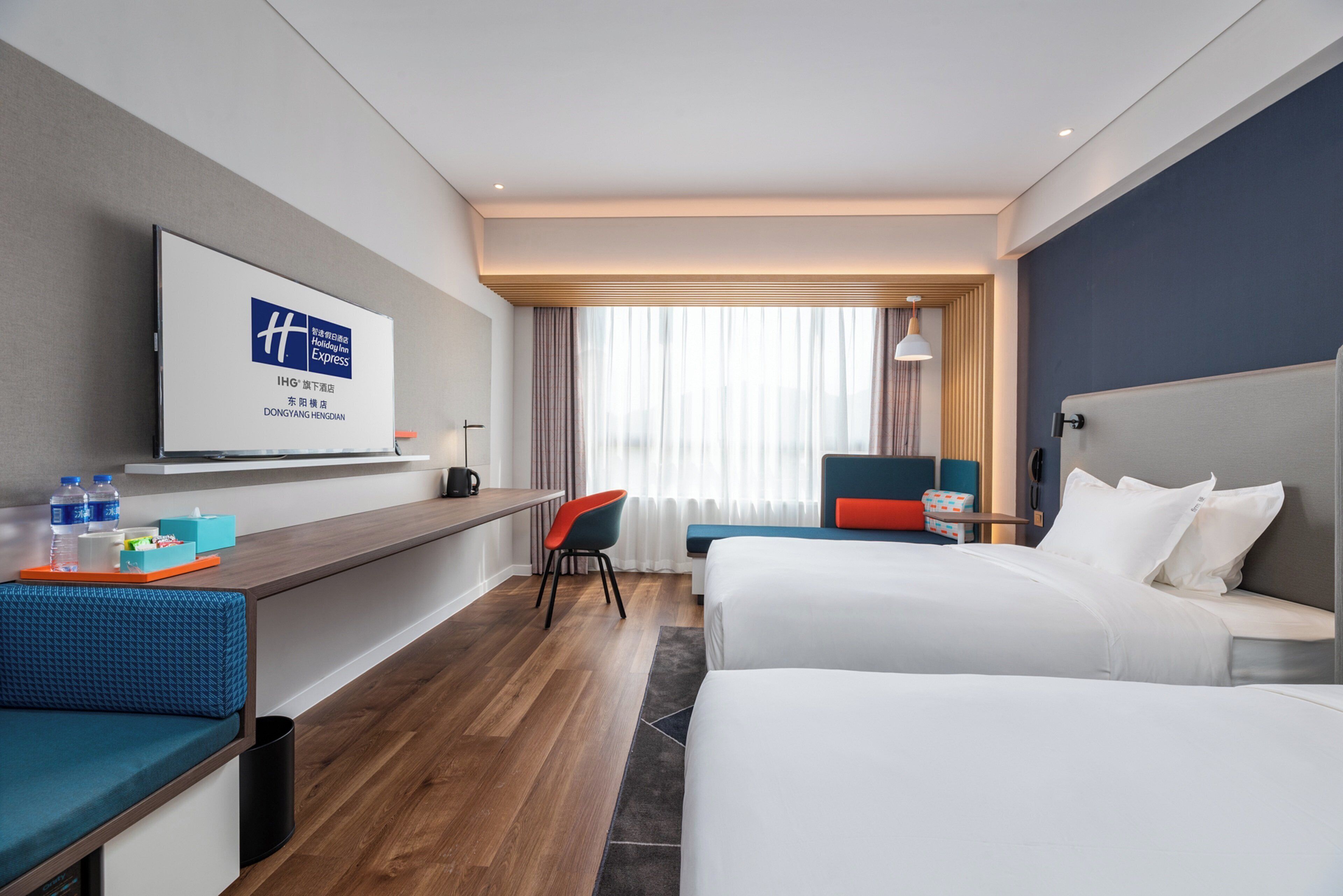 Ein Hotelzimmer mit Bett, Schreibtisch, Stuhl, Fernseher und einem fensterbeprägten Bereich mit Vorhängen.