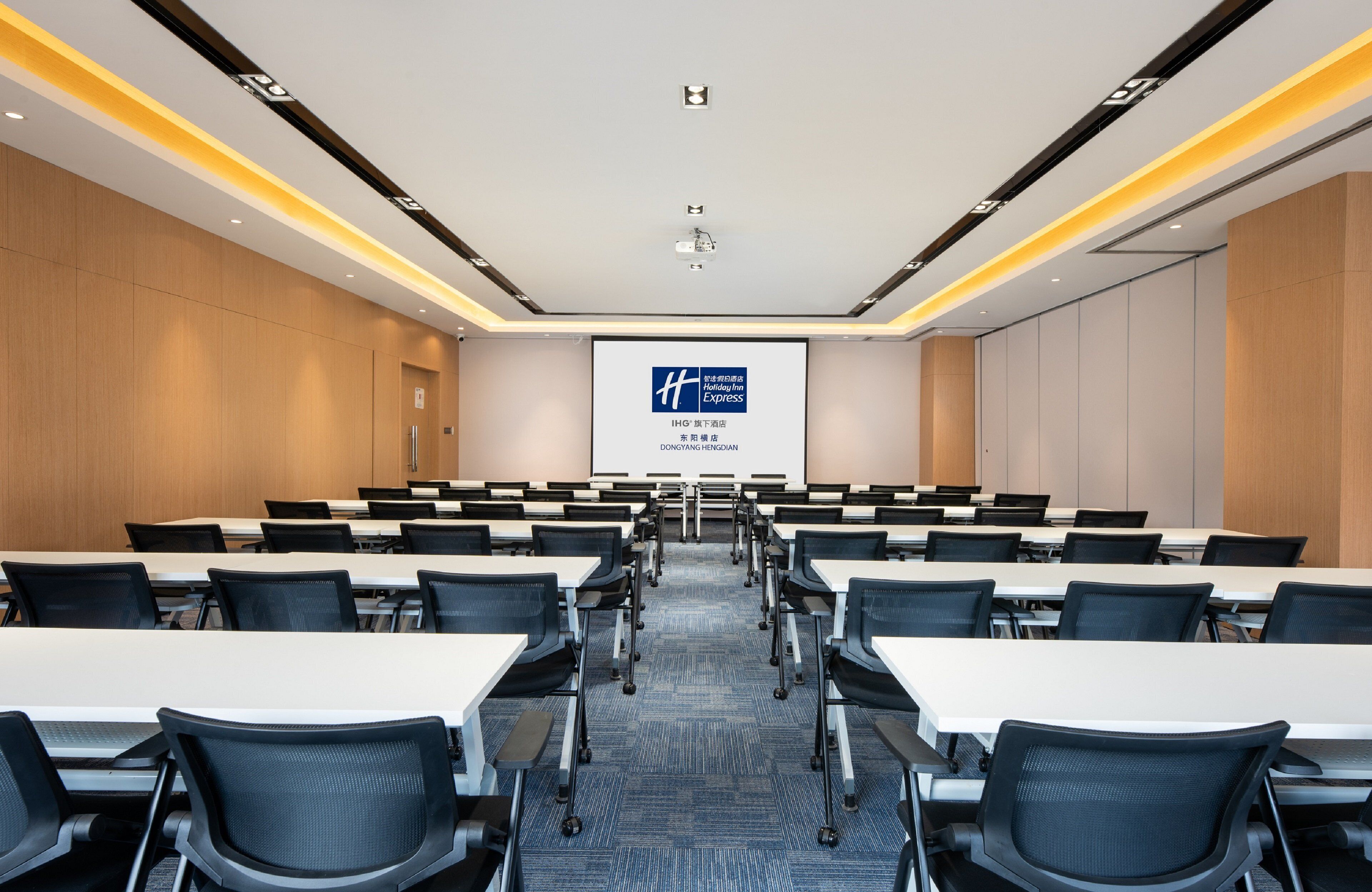 A conference room with rows of tables and chairs, a projector screen, and a presentation on the wall.