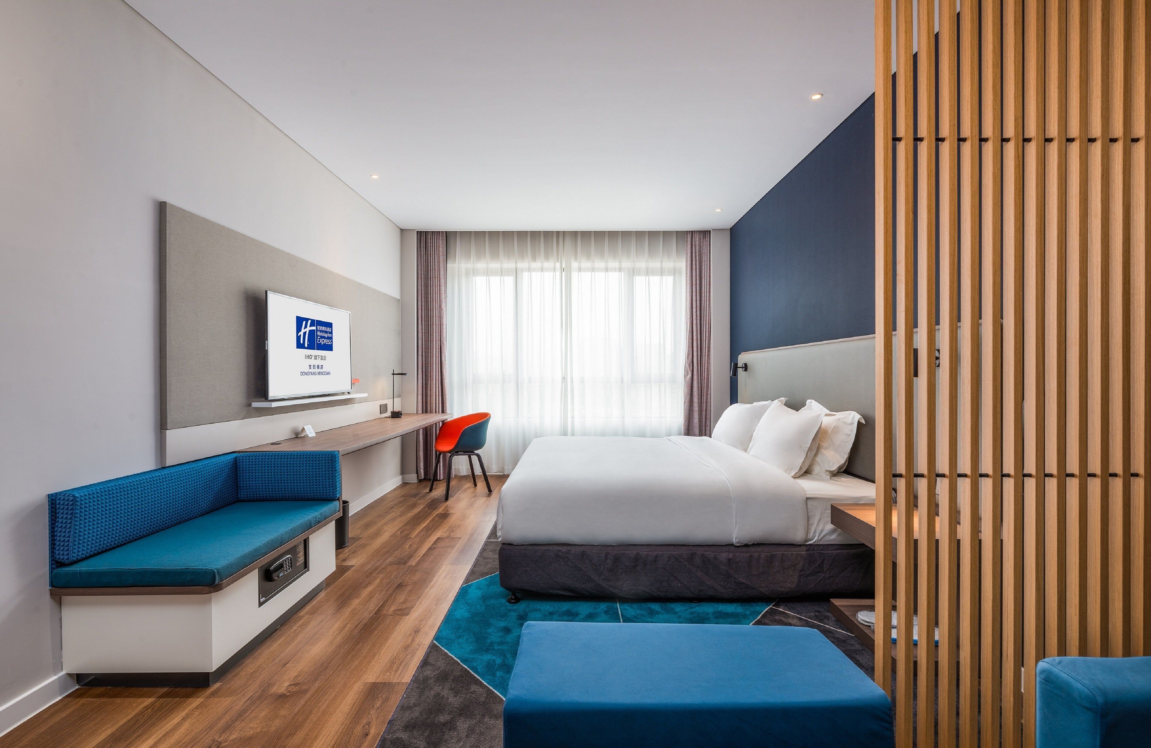 A modern hotel room with a bed, a seating area, a desk, and a TV on the wall.