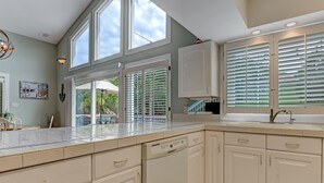 A kitchen with white cabinets, a large window with blinds, and a view of the outdoors. - Heated Pool Access, Great Outdoor Space (Tybee Island)