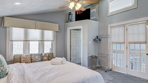 A bedroom with a large bed, a ceiling fan, a window with blinds, and a door leading to another room. - Heated Pool Access, Great Outdoor Space (Tybee Island)