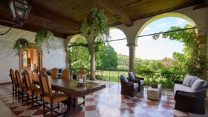 A covered patio with a checkered floor, a dining table with chairs, wicker furniture, hanging plants, and a view of the landscape. - Villa Torre di Terzolan With Pool and Jacuzzi (Verona)
