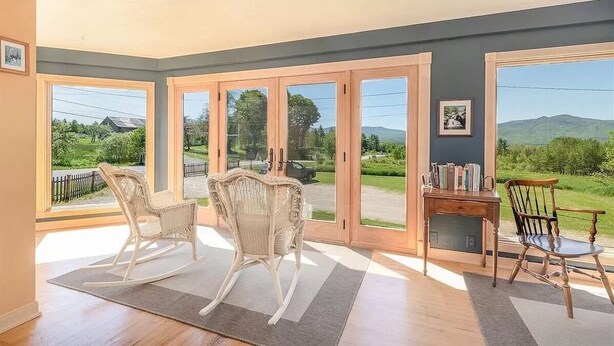 Quarto com grandes portas de vidro deslizantes, cadeiras de balanço de vime, uma mesa de madeira com livros e vista para áreas verdes e colinas.