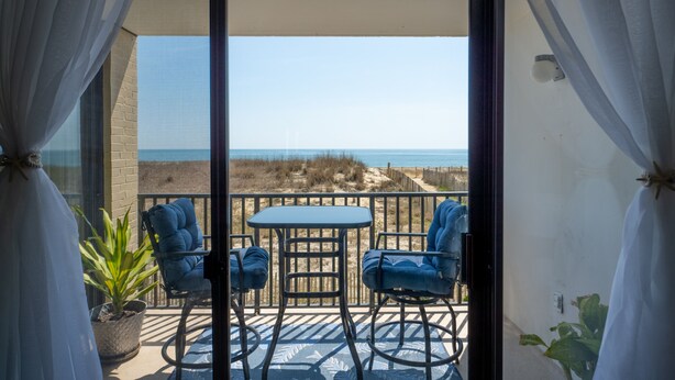 Un balcon avec une table et deux chaises, donnant sur une plage bordée de dunes et sur l’océan.