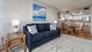 Un salon avec un canapé bleu, deux tables d’appoint avec des lampes, un coin repas avec des chaises en bois et, au fond, une cuisine.