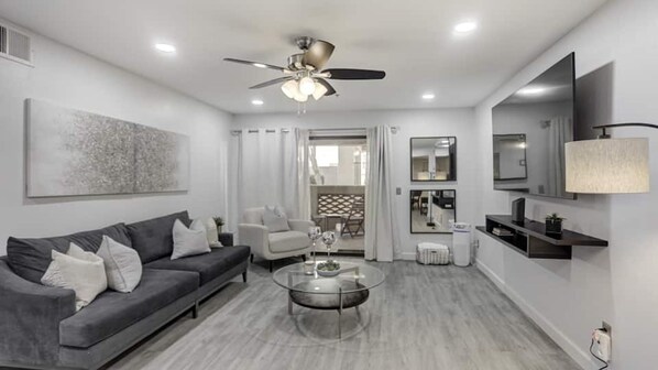 A modern living room with a grey sofa, a glass coffee table, a ceiling fan, and a view into the dining area. - Executive 2 Bdrm Condo in the Heart of Biltmore! (Phoenix)