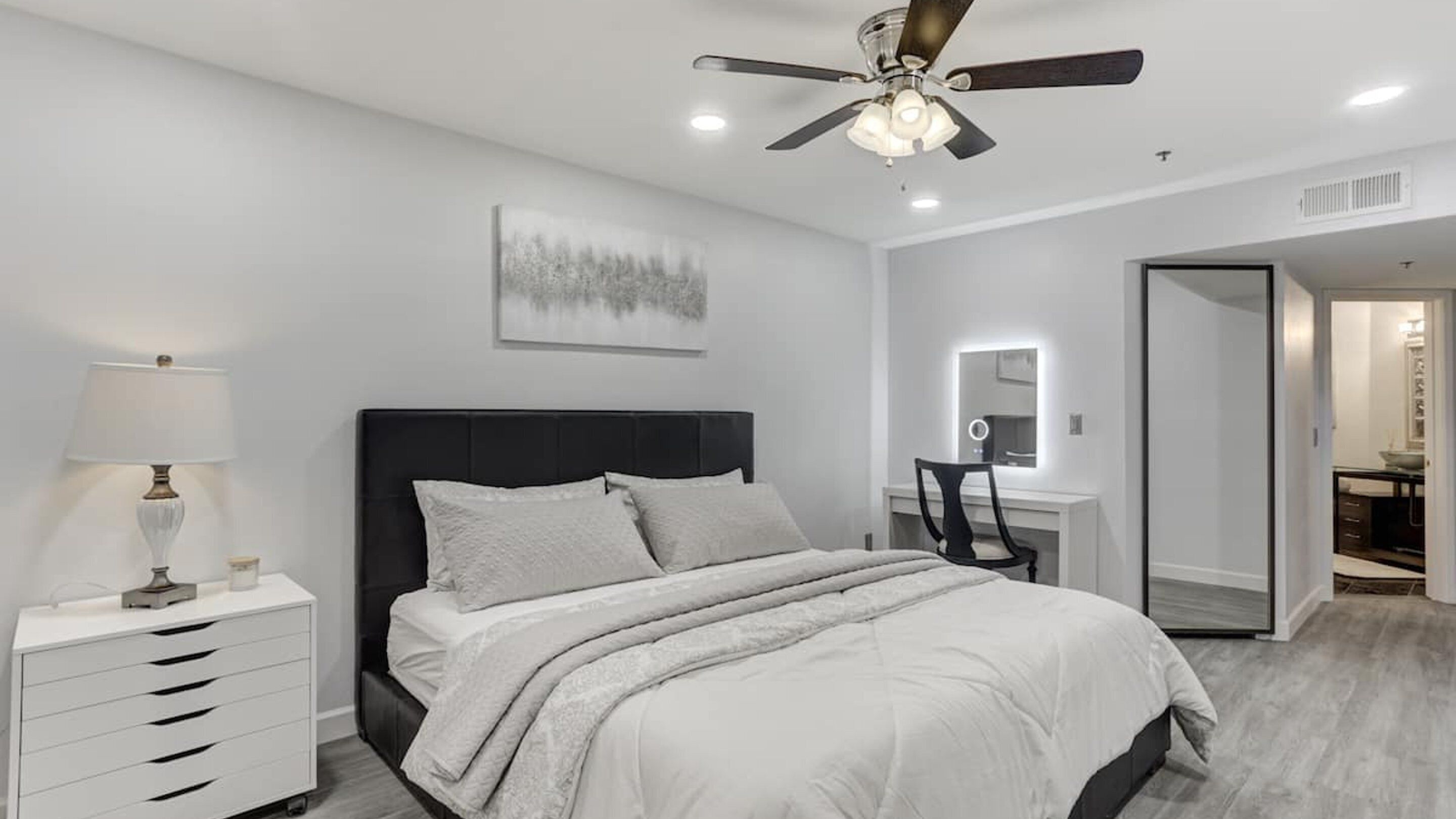 A modern bedroom with a large bed, a bedside table with a lamp, a ceiling fan, and a bathroom visible through an open door.