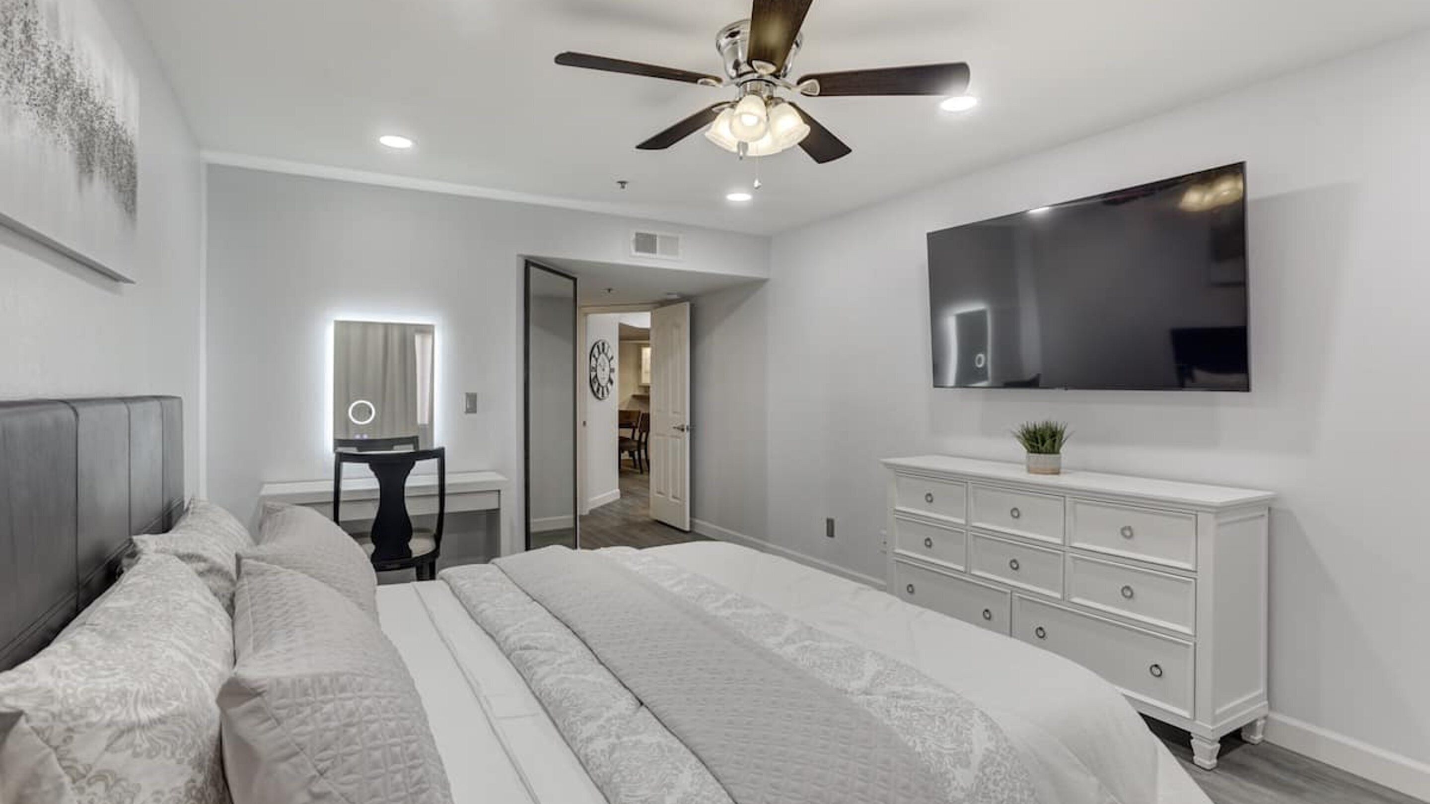 A bedroom with a bed, a dresser, a mirror, a ceiling fan, and a TV on the wall.