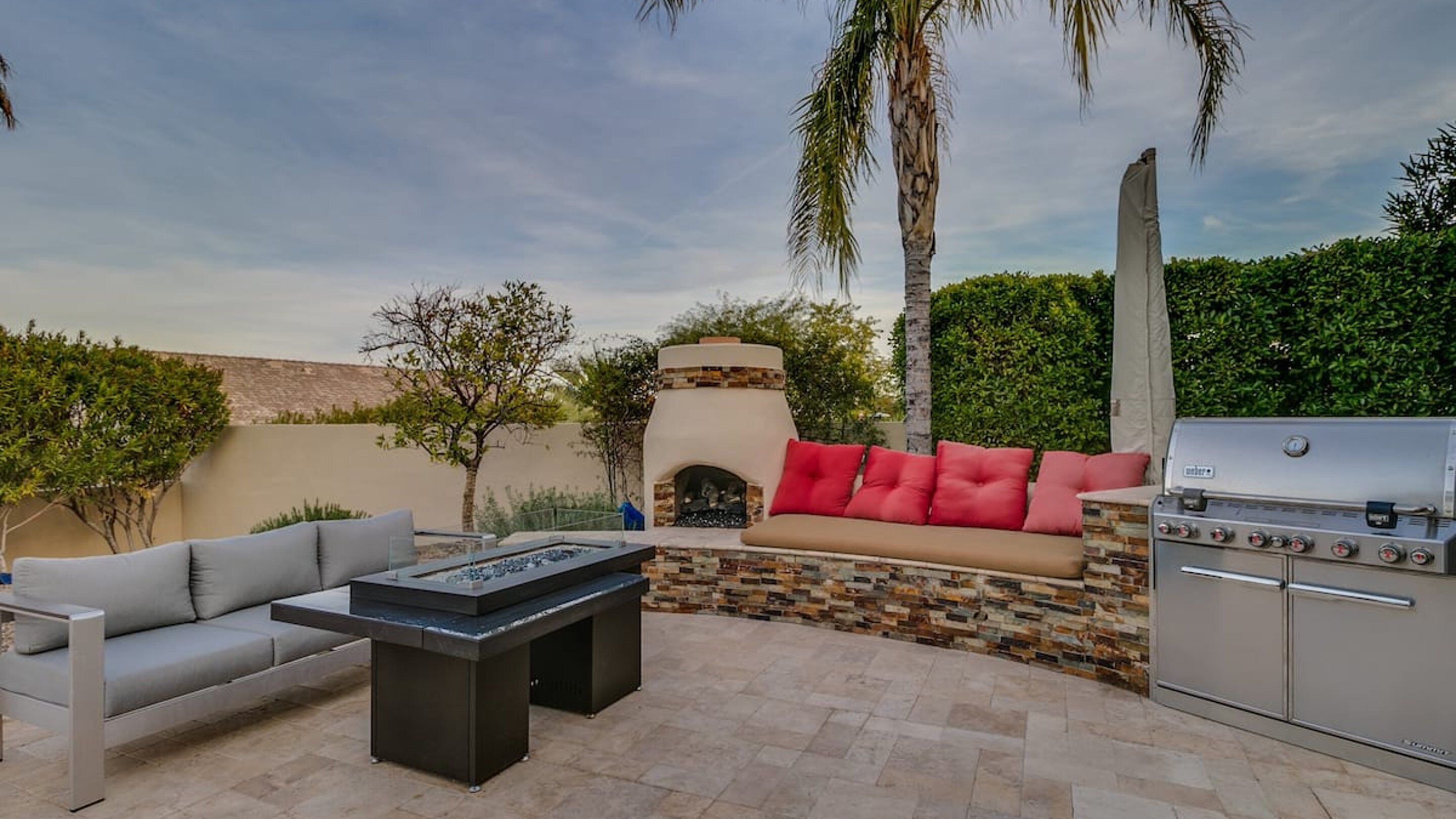 An outdoor patio with a stone fireplace, a grill, a sofa with red cushions, and a coffee table.