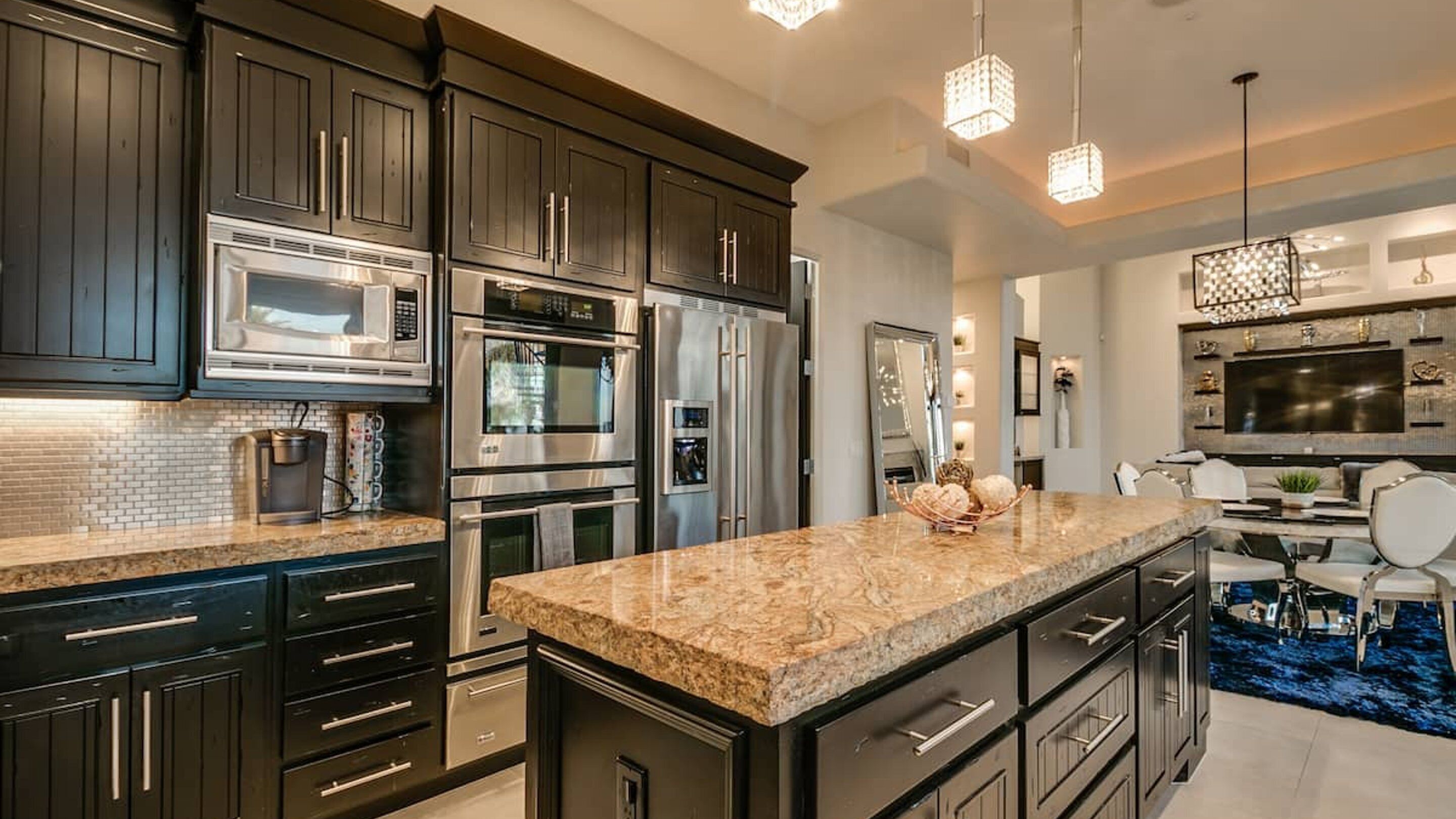 A modern kitchen with dark cabinetry, stainless steel appliances, a granite countertop, and a dining area with a blue rug.