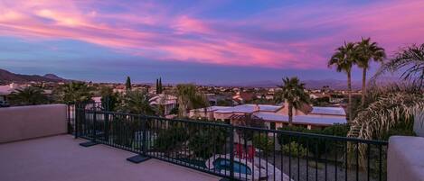 A rooftop terrace with a pool, palm trees, and a view of a residential area during sunset.