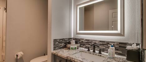 A bathroom with a white toilet, a sink with a granite countertop, a mirror with a light, and a towel rack.