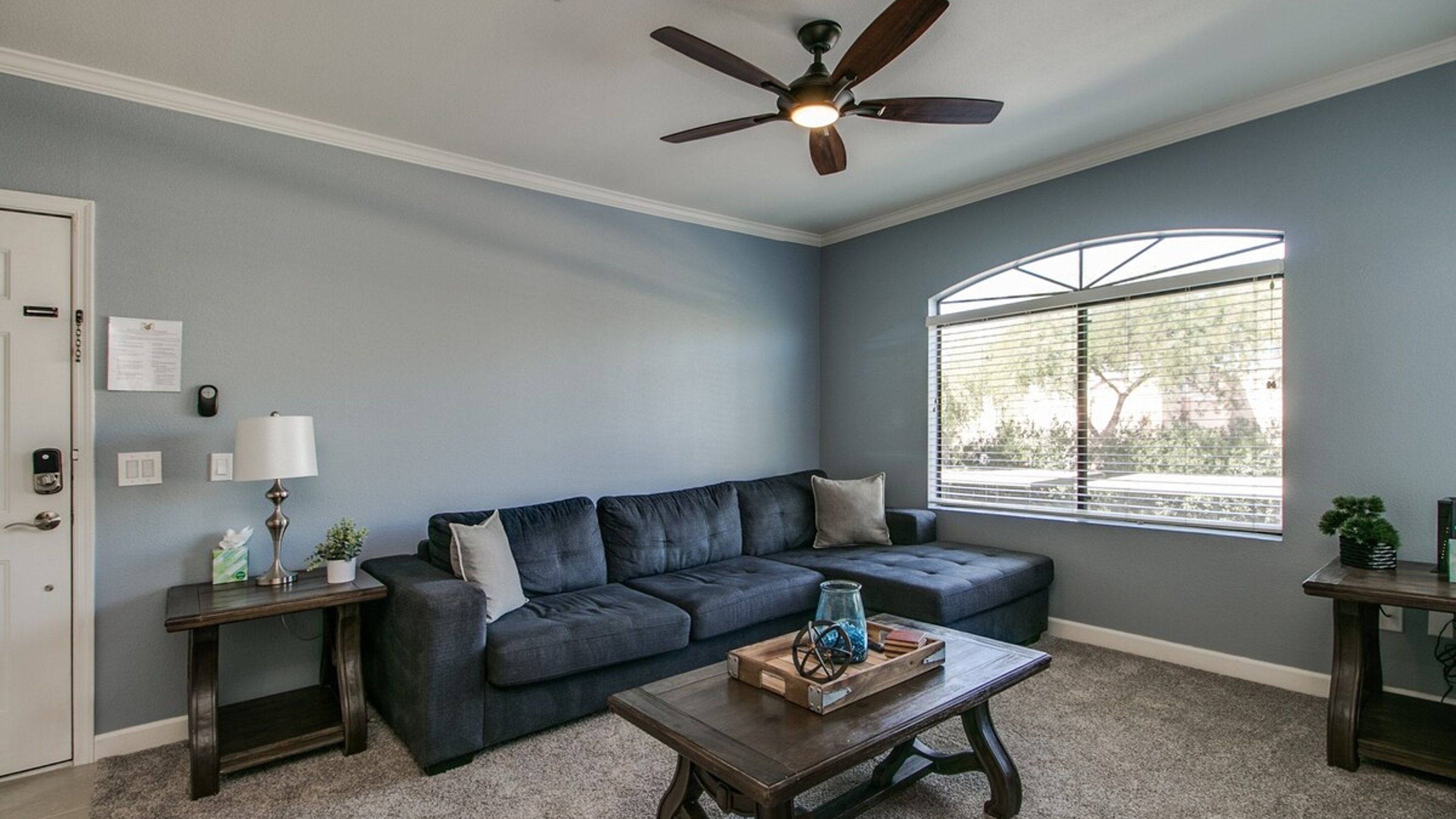 A living room with a dark blue sofa, a wooden coffee table, a ceiling fan, a window with blinds, and a door with a keypad lock.