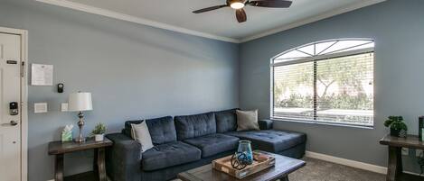 A living room with a dark blue sofa, a wooden coffee table, a ceiling fan, a window with blinds, and a door with a keypad lock.
